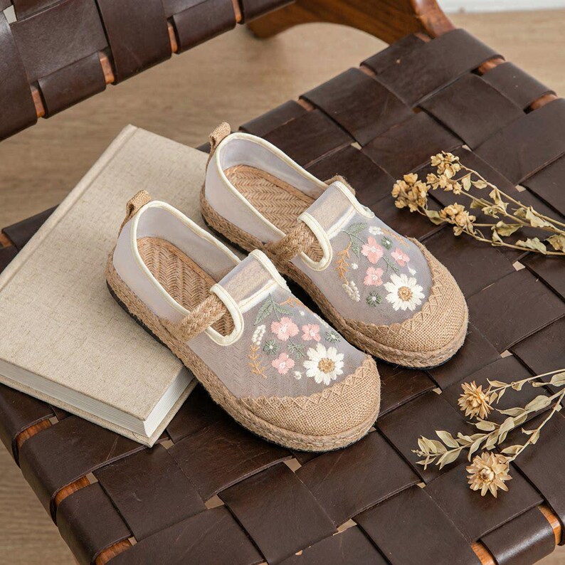 May include: A pair of beige and tan espadrille flats with floral embroidery. The shoes have a jute sole, a sheer overlay, and a strap closure. The flats are decorated with pink and white embroidered flowers.