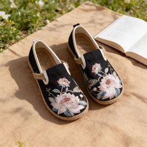 K&ouml;nnte beinhalten: Paar schwarze, bestickte Slipper mit floralen Mustern in Rosa, Blau und Gr&uuml;n. Die Schuhe haben eine Jutesohle und ein cremefarbenes Innenfutter. Im Hintergrund ist ein Buch ge&ouml;ffnet.