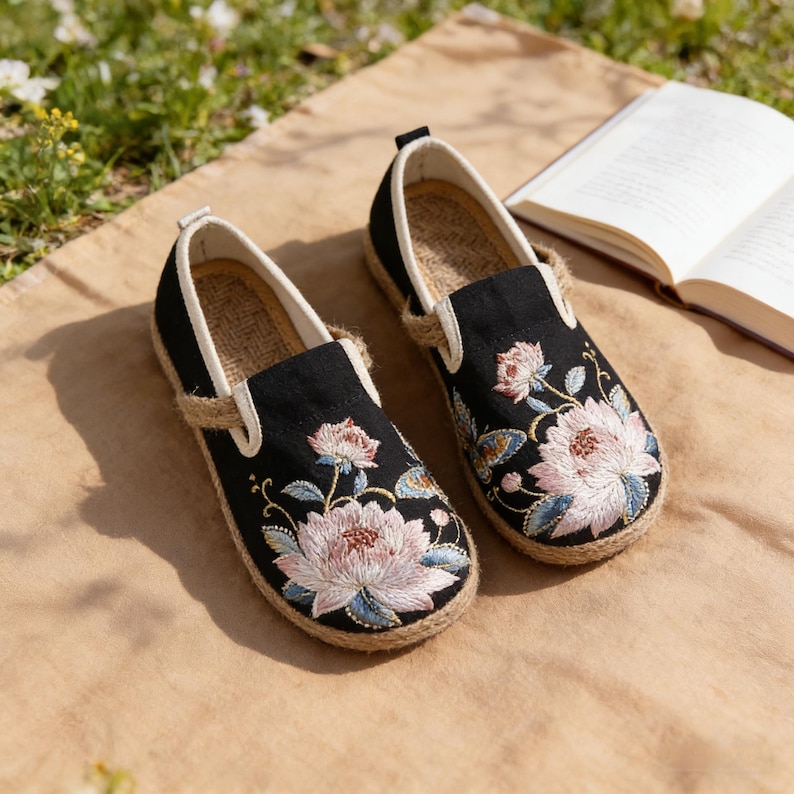 K&ouml;nnte beinhalten: Paar schwarze, bestickte Slipper mit floralen Mustern in Rosa, Blau und Gr&uuml;n. Die Schuhe haben eine Jutesohle und ein cremefarbenes Innenfutter. Im Hintergrund ist ein Buch ge&ouml;ffnet.