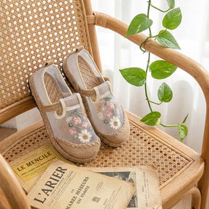 May include: A pair of beige espadrille shoes with floral embroidery. The shoes have a woven jute sole and a sheer overlay on the top, featuring pink and white flowers. They are placed on a woven chair with vintage papers.