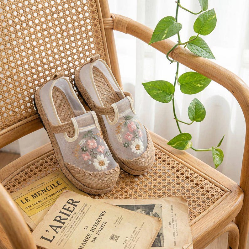 May include: A pair of beige espadrille shoes with floral embroidery. The shoes have a woven jute sole and a sheer overlay on the top, featuring pink and white flowers. They are placed on a woven chair with vintage papers.