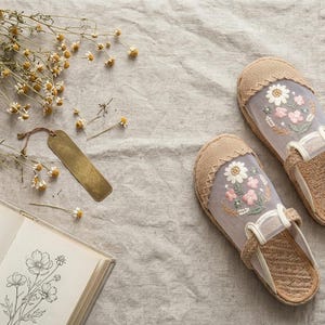 May include: Embroidered floral slippers in beige and white, with a sheer overlay. The slippers are on a light beige linen surface, next to a book with floral illustrations and dried flowers. A brass bookmark is also present.
