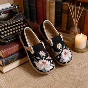 K&ouml;nnte beinhalten: Paar schwarze Slipper mit floraler Stickerei in Rosa, Wei&szlig; und Blau. Die Schuhe haben eine Juteseil-Umrandung und eine Innensohle aus Naturfaser. Das Design zeichnet sich durch einen klassischen, bequemen Stil aus.