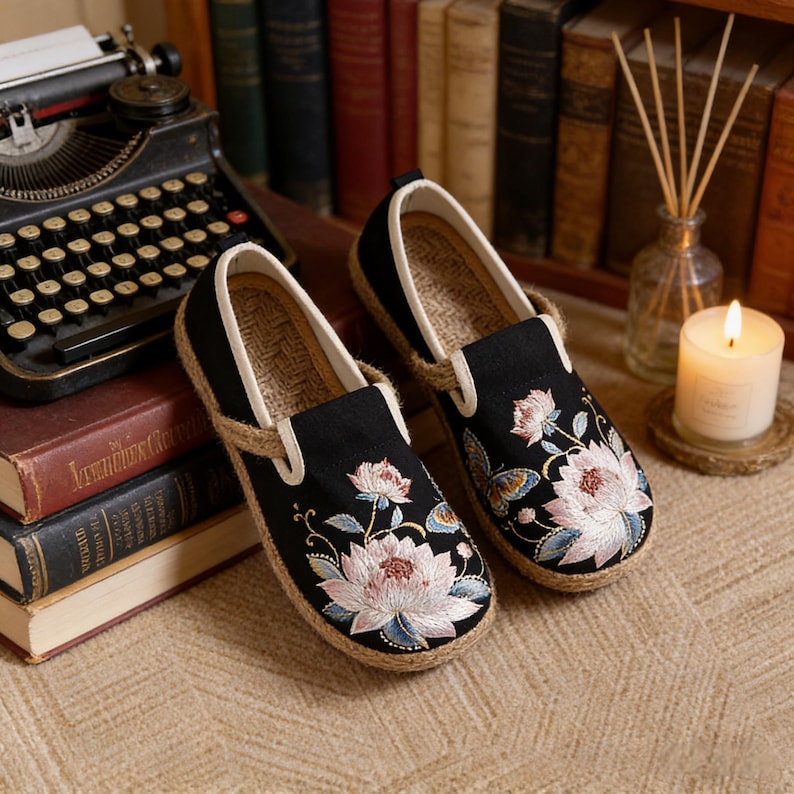 K&ouml;nnte beinhalten: Paar schwarze Slipper mit floraler Stickerei in Rosa, Wei&szlig; und Blau. Die Schuhe haben eine Juteseil-Umrandung und eine Innensohle aus Naturfaser. Das Design zeichnet sich durch einen klassischen, bequemen Stil aus.