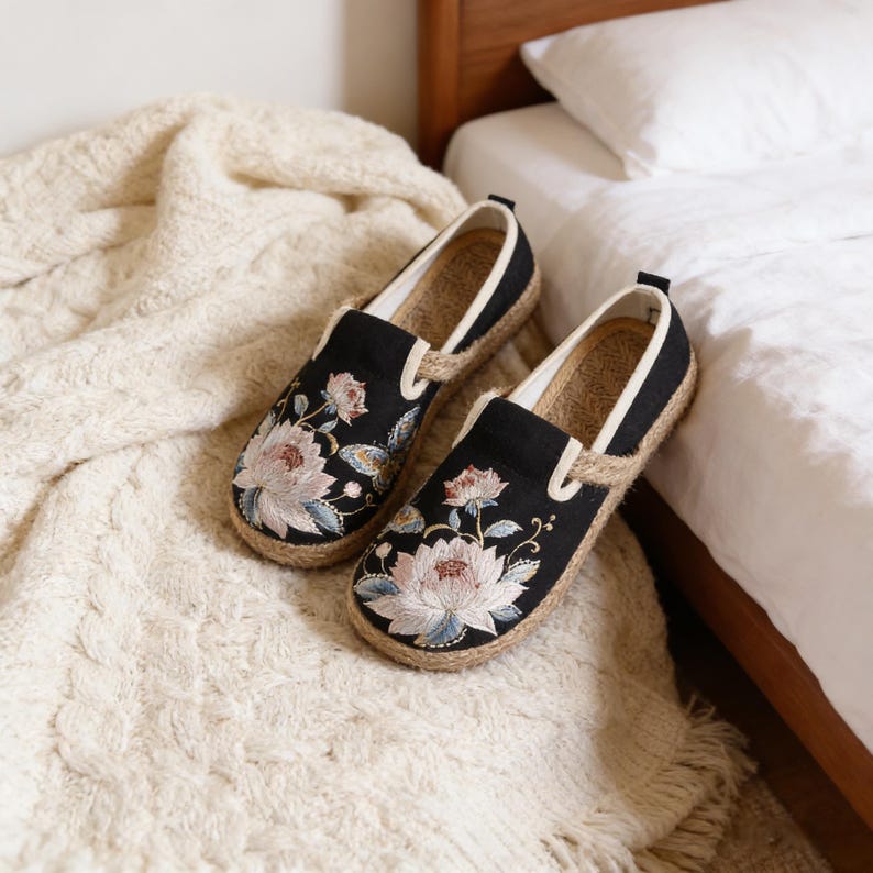 K&ouml;nnte beinhalten: Schwarze Slipper mit floraler Stickerei. Die Schuhe haben eine Jutesohle und einen wei&szlig;en Besatz. Die gestickten Blumen sind in Rosa, Blau und Wei&szlig; gehalten. Die Schuhe liegen auf einer cremefarbenen Decke.
