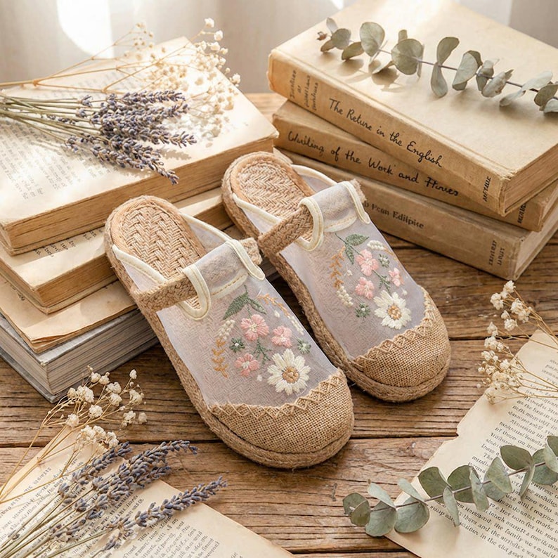 May include: Embroidered slippers in beige and tan, featuring floral patterns. The slippers have a woven base and a sheer overlay with pink and white flowers. The background includes books and dried lavender.