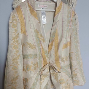 May include: A cream-colored blazer by Christian Lacroix featuring a textured fabric with yellow and light blue floral patterns, vertical stripes, and a front tie closure. The garment has long sleeves and a label indicating size 40 at the neckline.