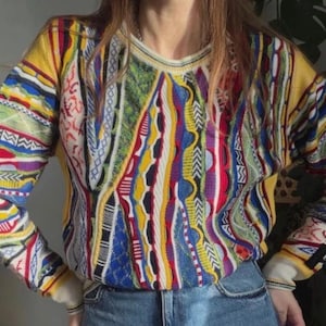 Pode incluir: Uma camisola de manga comprida com gola redonda e um padrão de malha complexo e multicolorido. O design incorpora tons vibrantes de amarelo, azul, vermelho, verde e branco em várias riscas verticais e horizontais geométricas e texturizadas.