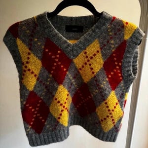 May include: A sleeveless V-neck sweater vest featuring a gray, red, and yellow argyle diamond pattern. The knit garment has a ribbed neckline, armholes, and hem, with a black label visible at the inner collar area. The texture appears fuzzy.