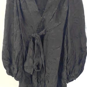 May include: A black long-sleeve blouse featuring a V-neckline, a front tie closure, and gathered puff sleeves with elasticized cuffs. The fabric has a subtle tonal jacquard pattern throughout, creating a textured appearance on the garment surface.