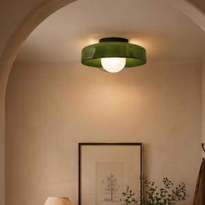 May include: A green and black flush mount ceiling light with a white globe bulb. The light fixture is mounted in a hallway with a neutral color palette. A framed picture, lamp, and vase are visible.