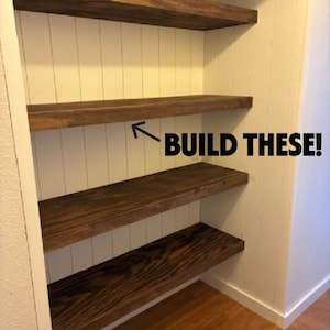 May include: A built-in shelving unit with three dark stained wooden shelves against a white paneled wall. The text "BUILD THESE!" is visible on the wall. The shelves are supported by the white wall and are above a wooden floor.