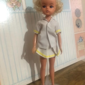 以下が含まれることがあります： ブロンドの髪と青い目のヴィンテージドール。白いテニスウェアに黄色のトリムが付いています。透明なスタンドに立ち、白いスニーカーを履いています。背景には装飾的な壁パネルがあります。