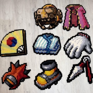 Op de afbeelding: Een verzameling pixel art tapijten in diverse vormen, waaronder een waaier met een cirkelvormig ontwerp, een bruine helm, een roze en rode cape, een blauw-witte bundel, een witte handschoen met rode accenten, een rood stekelig object, een geel-grijze laars en een wit-rode dolk.