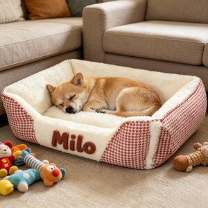 Puede incluir: Una cama rectangular para mascotas con interior de felpa color crema y exterior con estampado de pata de gallo en rojo y blanco. El nombre Milo está bordado en letras marrones en la parte frontal. Un perro pequeño beige duerme dentro.