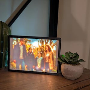 May include: A rectangular digital photo frame with a black border displaying a backlit image of a family of four standing outdoors in a wooded area during autumn. The frame is positioned between a tall green ceramic cactus and a small succulent plant.