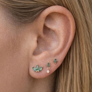 May include: Close-up of an ear adorned with four gold earrings. The earrings feature green gemstones in various shapes, including a cluster, a teardrop, a diamond, and a dangling design with a small diamond accent. The earrings are set in gold.