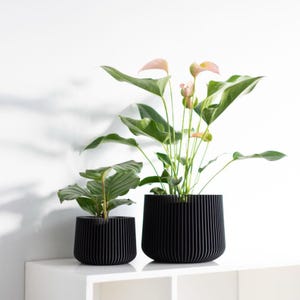 Op de afbeelding: Twee zwarte, geribbelde plantenbakken met groene planten en roze bloemen. De plantenbakken zijn verschillend van grootte, waarbij de grotere een hogere plant bevat. De plantenbakken staan op een witte plank tegen een witte muur.