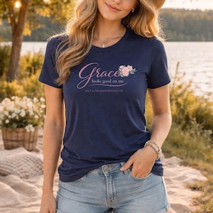 May include: Navy blue t-shirt with the text "Grace looks good on me" in pink script and a floral design. The shirt is from Salt & Breaker Revival Co. The model is wearing a straw hat and denim shorts.