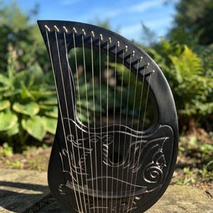 May include: A black wooden lyre featuring a carved dragon design on its body. The instrument has a curved frame with multiple thin metal strings stretched vertically across the open sound box, secured by small metal tuning pins along the top edge.