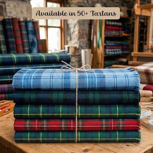 May include: A stack of five folded tartan blankets tied with twine, featuring blue, green, and red plaid patterns with fringed edges. The text "Available in 50+ Tartans" is visible at the top of the image, indicating a variety of color options.