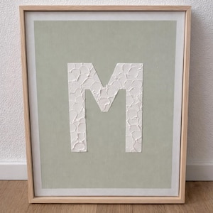 May include: A framed artwork featuring the letter "M" in white, textured paint against a muted green background. The artwork is encased in a light-colored wooden frame, creating a minimalist and modern aesthetic. The piece is displayed on a wooden floor.