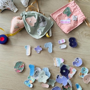 Puede incluir: Una colección de números y formas de tela hechos a mano en varios colores y patrones, incluyendo diseños florales y a rayas. La imagen también presenta dos bolsas con cordón, una cinta métrica y artículos de tela en forma de corazón y estrella.