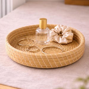 May include: A round woven tray holds a clear perfume bottle with a gold cap, a beige satin scrunchie, and gold hoop earrings and a bracelet. The tray is made of light brown woven material.