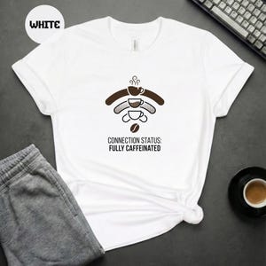 Op de afbeelding: Wit T-shirt met een koffiethema. Het ontwerp toont een koffiekopje-pictogram in een Wi-Fi-symbool, met de tekst "CONNECTION STATUS: FULLY CAFFEINATED" eronder. Het shirt wordt gecombineerd met een grijze joggingbroek en een toetsenbord.