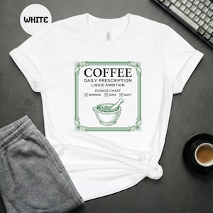 Op de afbeelding: Wit T-shirt met een groen en witte afbeelding. De afbeelding luidt "COFFEE DAILY PRESCRIPTION LIQUID AMBITION DOSAGE CHART" met vinkjes voor ochtend, middag en nacht. Een illustratie van een vijzel en stamper is ook inbegrepen.