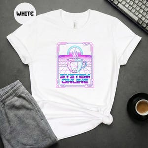 Op de afbeelding: Wit T-shirt met een grafisch ontwerp met een geometrische koffiekop met stoom, tegen een retro rasterachtergrond. De tekst "SYSTEM ONLINE" staat eronder in een levendig, neon-achtig lettertype.