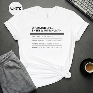 Op de afbeelding: Wit T-shirt met de tekst "OPERATOR SPEC SHEET // UNIT: HUMAN". De tekst bevat details over brandstoftype, huidige status, vereiste dosering en een waarschuwing. Een kopje koffie en een toetsenbord zijn ook zichtbaar.