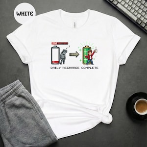 Op de afbeelding: Wit T-shirt met een afbeelding van een lege batterij die verandert in een volle batterij, met de tekst "DAILY RECHARGE COMPLETE". Het ontwerp is pixelig en toont een persoon met een koffiekopje. Het woord "WHITE" staat in een witte cirkel.