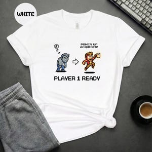 Op de afbeelding: Wit T-shirt met een pixelachtige afbeelding die een personage afbeeldt dat transformeert van een zombie in een energieke figuur met een mok. De tekst luidt "POWER UP ACQUIRED!" en "PLAYER 1 READY".