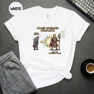 Op de afbeelding: Wit T-shirt met de tekst "CLASS UPGRADE COMPLETE". Het ontwerp toont een pixelige illustratie van een transformatie van een mens in een barista-magiër. Het T-shirt heeft een ronde hals en korte mouwen.