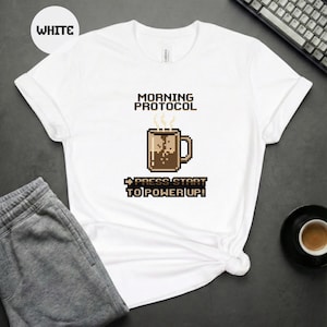 Könnte beinhalten: Weißes T-Shirt mit dem Text "MORNING PROTOCOL" über einer pixeligen Kaffeetasse. Unter der Tasse steht der Text "PRESS START TO POWER UP!". Das Shirt wird mit grauer Jogginghose und einer Tasse Kaffee kombiniert.