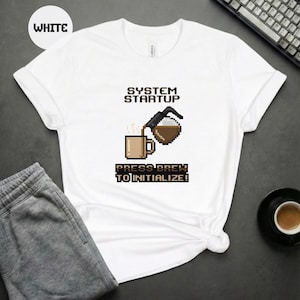 Op de afbeelding: Wit T-shirt met een pixelafbeelding met koffiethema. Het ontwerp toont een koffiepot die in een mok giet, met de tekst "SYSTEM STARTUP" erboven en "PRESS BREW TO INITIALIZE!" eronder. Het shirt ligt op een donkere ondergrond.