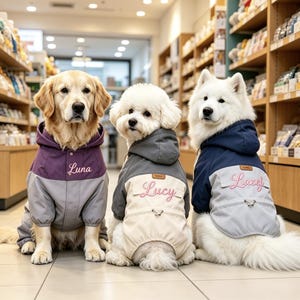 以下が含まれることがあります： お揃いの犬用ジャケットを着た3匹の犬。ジャケットはグレー、紫、ネイビーで、ライトグレーとクリーム色のパネルが付いています。各ジャケットの背面には名前が刺繍されています：ルナ、ルーシー、ラゼル。犬たちはペットショップにいます。