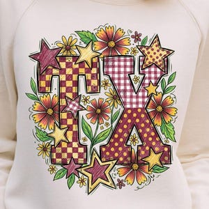 Peut inclure: Un sweat-shirt à manches longues de couleur crème arborant un motif central avec les lettres TX dans un style à carreaux et vichy. Le design est entouré d'illustrations colorées de fleurs variées et d'étoiles dans des tons jaune, rouge et vert.