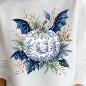 May include: A white crewneck sweatshirt featuring a central graphic of a pumpkin decorated with a blue and white toile pattern. The pumpkin is surrounded by dark blue bat wings and various botanical elements including leaves and small sprigs.