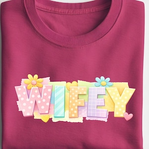 May include: A maroon crew neck t-shirt featuring the word "WIFEY" in a colourful, decorative font. The letters are decorated with flowers, hearts, and polka dots. The t-shirt is neatly folded.