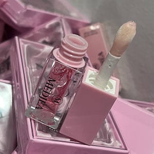 May include: A clear lip gloss tube with a pink cap and applicator. The gloss is a light pink color and the tube has the word "MEDJEA" printed on it. The lip gloss is packaged in a pink box with the number "018" printed on it.