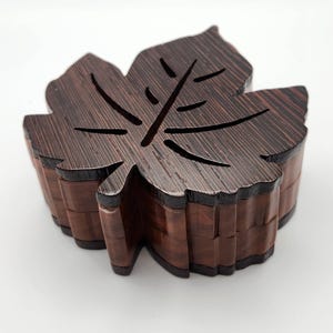 Könnte beinhalten: Eine holzfarbene Schatulle in Blattform mit dunkelbraunem Finish und sichtbarer Holzmaserung. Die Schatulle hat ein geschichtetes Design mit einem geschnitzten Blattmuster auf dem Deckel und einem kontrastierenden dunklen Rand.