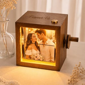 May include: A square wooden box with a dark brown finish featuring the engraved text Forever Us on the top. The front displays an illuminated photograph of a couple. A small brass button sits on the top, and a wooden crank handle is on the side.