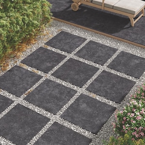Puede incluir: Diseño de patio exterior con baldosas cuadradas de color gris oscuro colocadas en un lecho de grava. Las baldosas están dispuestas en un patrón de cuadrícula, rodeadas de vegetación y una tumbona de madera.