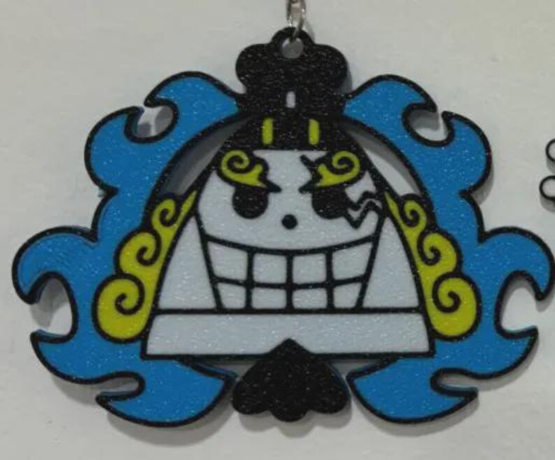 May include: A keychain featuring a stylized, grinning skull with a black top, blue wavy accents, and yellow details. The skull has a white face with black outlines and a large, toothy grin. The keychain is hanging from a silver chain.