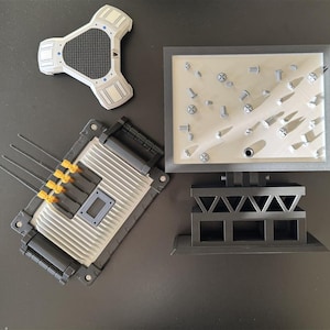 Può includere: Una collezione di componenti di modelli grigi e neri, tra cui un pezzo triangolare bianco e grigio, un pezzo rettangolare con antenne e una base rettangolare con viti. I pezzi sono disposti su una superficie scura.