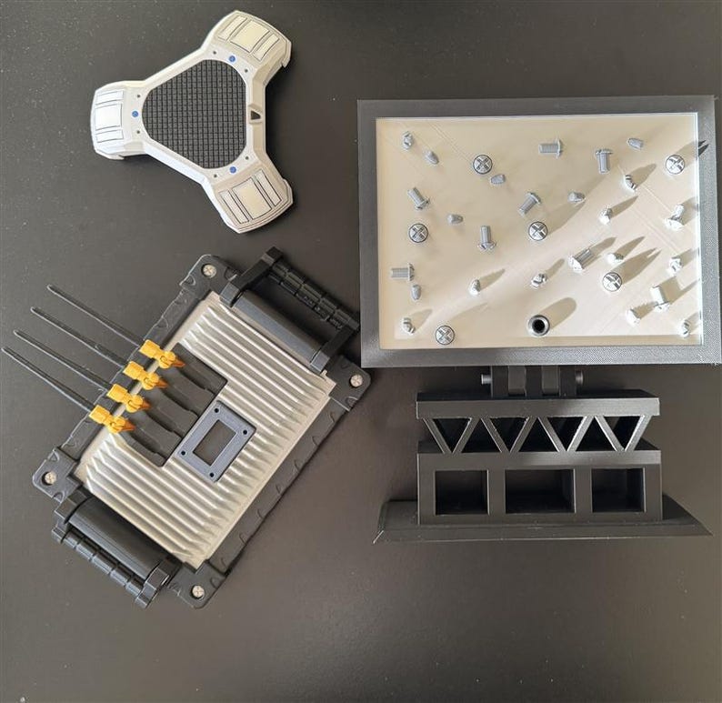 May include: A collection of gray and black model components, including a triangular white and gray piece, a rectangular piece with antennas, and a rectangular base with screws. The pieces are arranged on a dark surface.