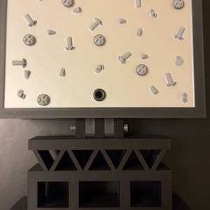 May include: A gray 3D-printed display featuring various screws and bolts arranged on a light beige surface. The display is mounted on a dark gray, geometric stand. The screws and bolts are in various sizes and orientations.
