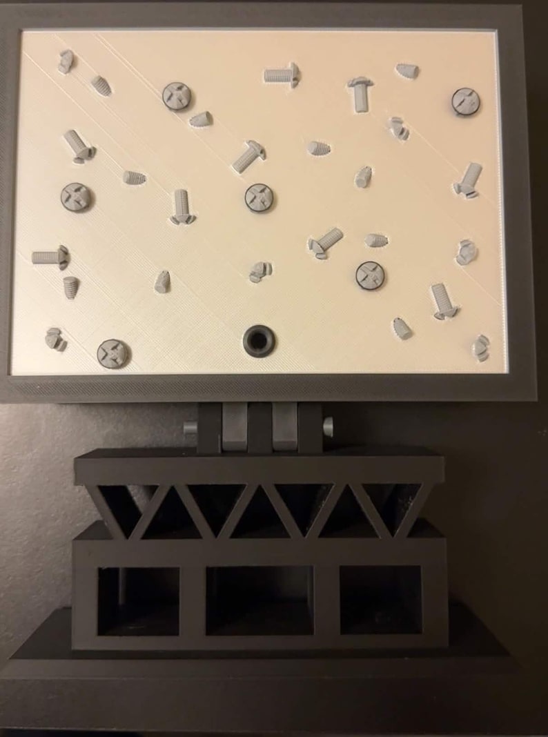 May include: A gray 3D-printed display featuring various screws and bolts arranged on a light beige surface. The display is mounted on a dark gray, geometric stand. The screws and bolts are in various sizes and orientations.